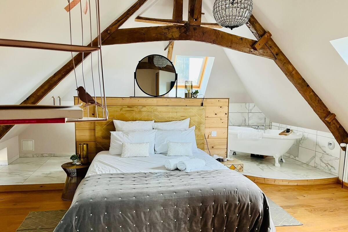 A bedroom with a large bed, a wooden headboard, a round mirror, a hanging light fixture, and a view of the bathroom.