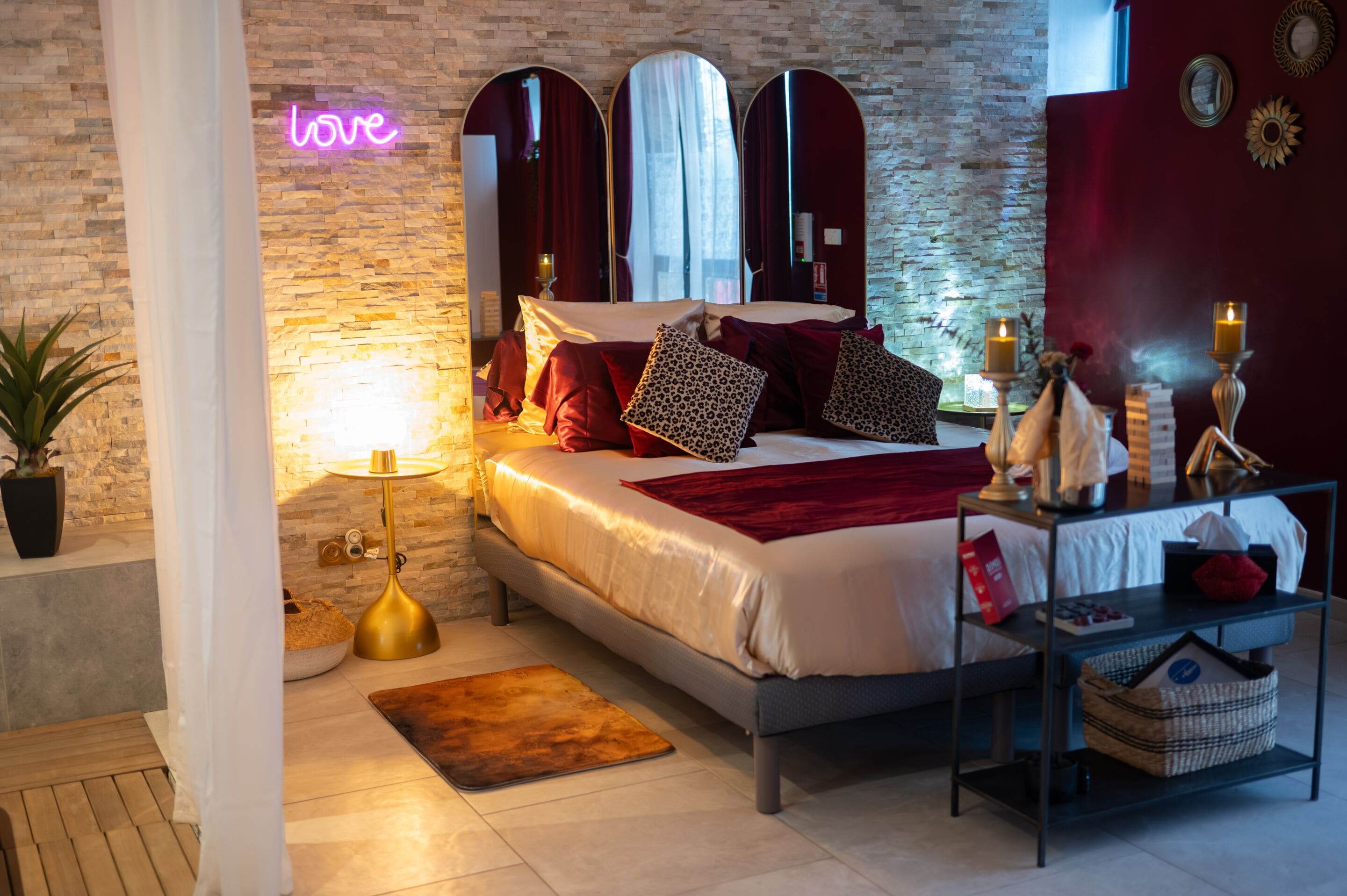 A modern bedroom with a brick wall, a neon 'love' sign, a bed with red and patterned pillows, a bedside table with candles, and a small plant.
