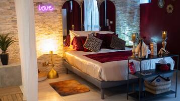A modern bedroom with a brick wall, a neon 'love' sign, a bed with red and patterned pillows, a bedside table with candles, and a small plant.