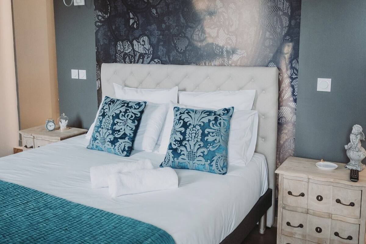 A neatly made bed with a white and blue patterned duvet, a teal throw blanket, and matching pillows. There is a bedside table with a lamp and a small decorative item. The room has a dark patterned wallpaper and a light-colored headboard.