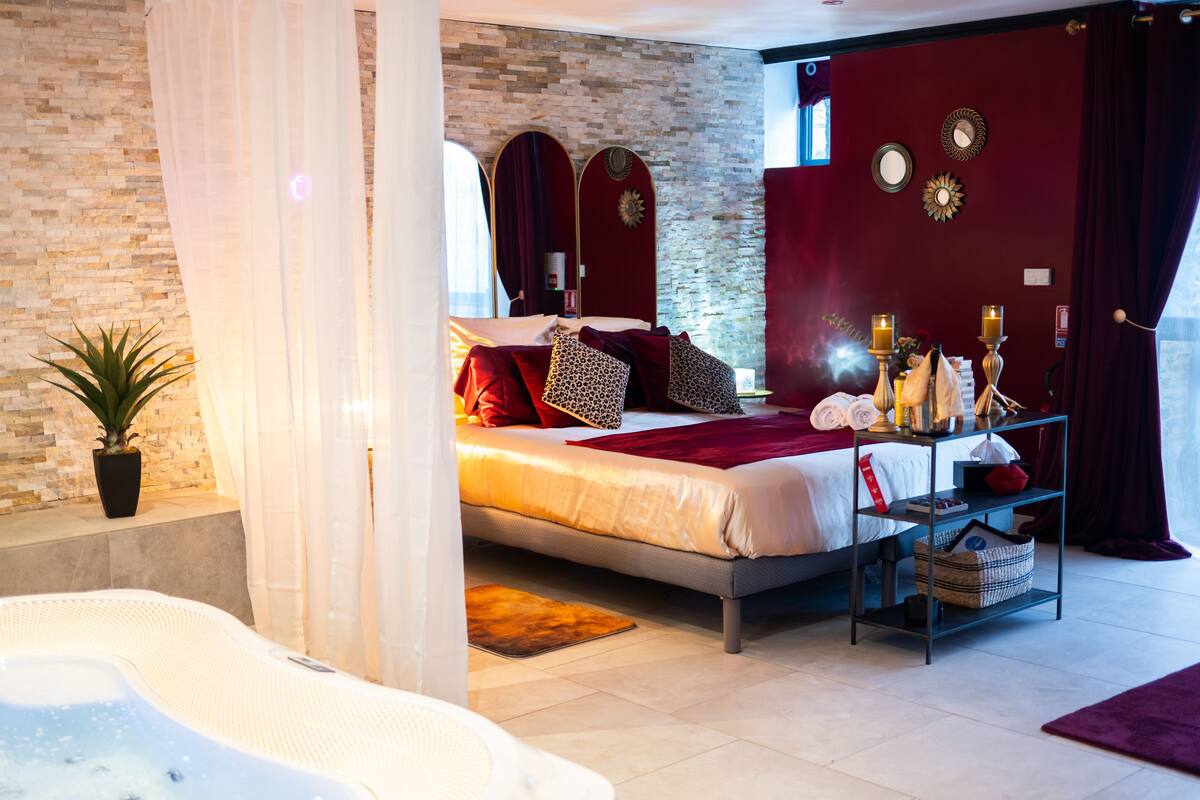 A modern bedroom with a large bed, a bedside table with candles, a small table with a basket, and a hot tub in the foreground.