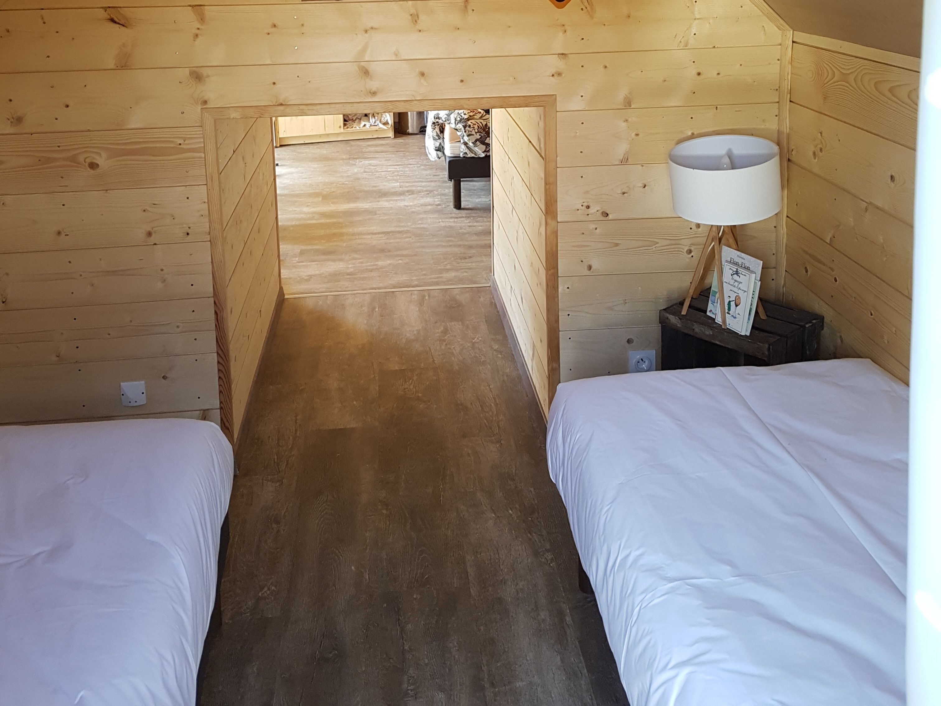 A wooden cabin room with two beds, a lamp, and a small table with books.