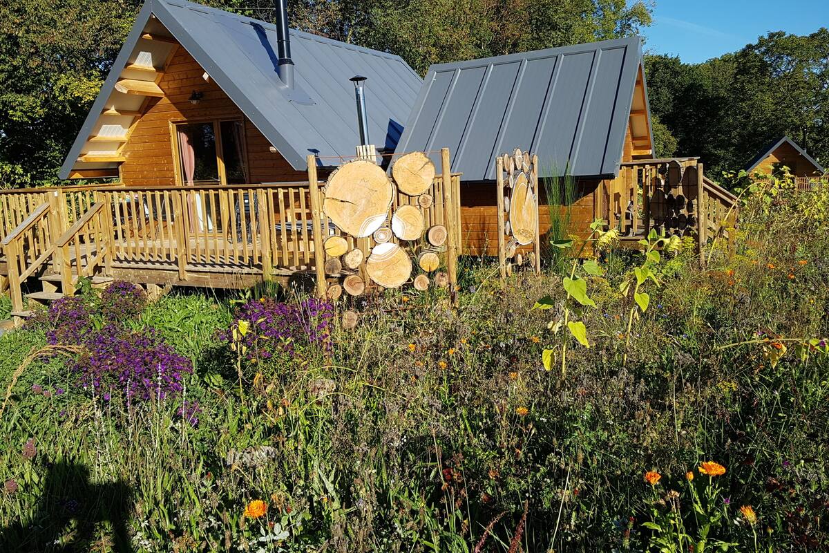 A wooden cabin with a sloped roof, a wooden deck, and a stack of logs.