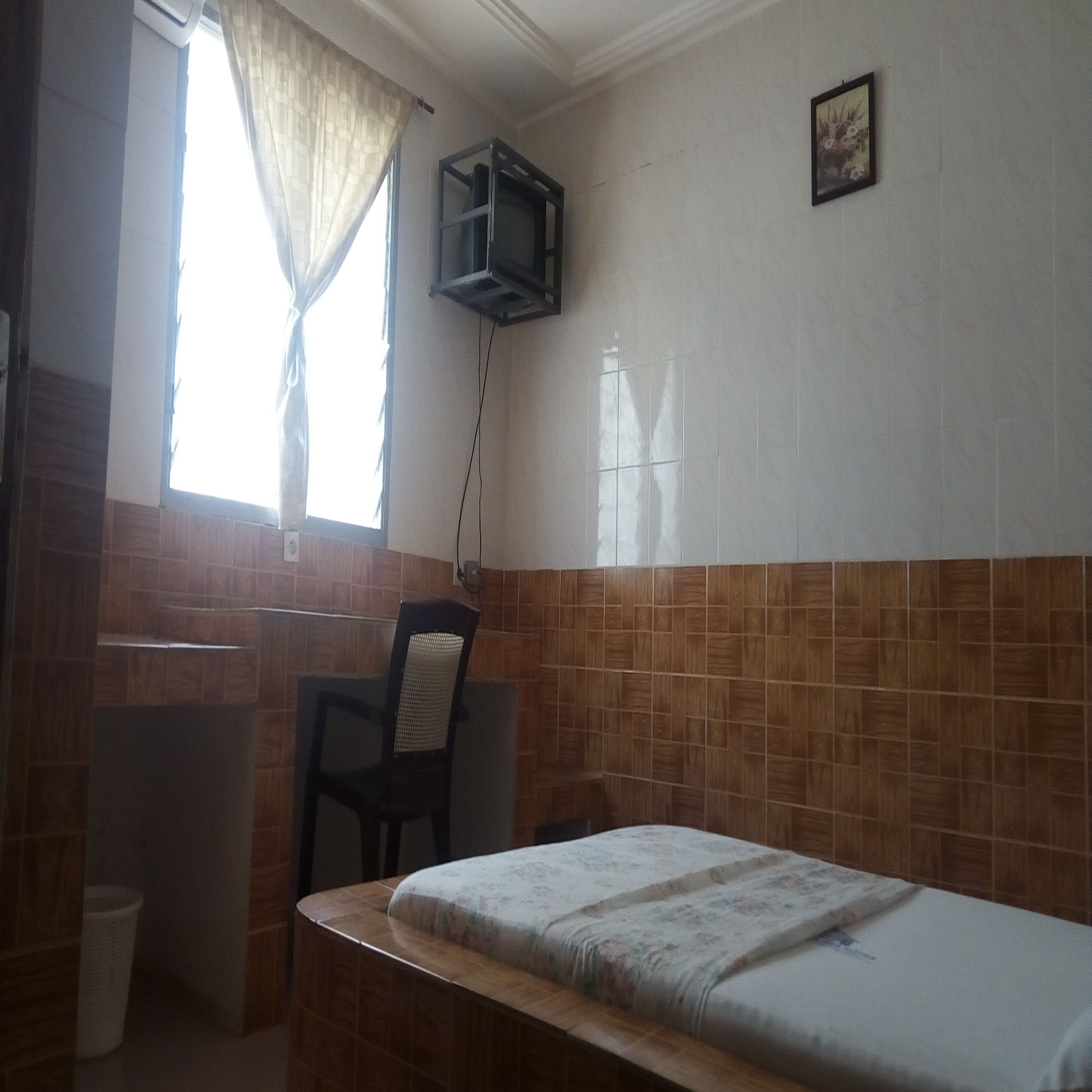 Una habitación pequeña y sencilla con cama, silla, ventana con cortinas y estante en la pared.