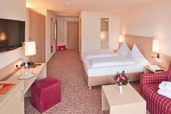 A hotel room with a bed, a TV, a desk with a lamp, a red ottoman, and a red sofa.