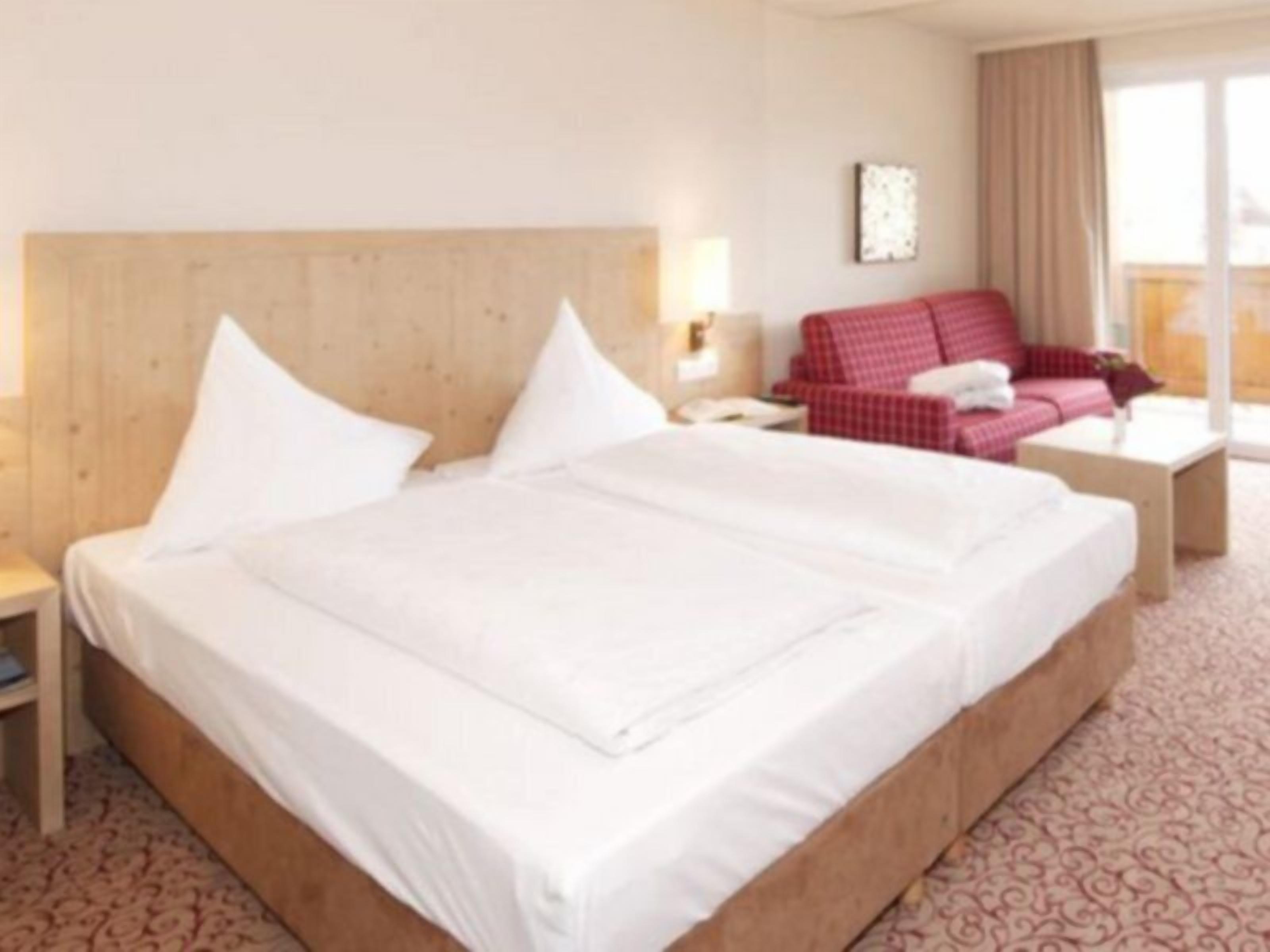 A hotel room with a large bed, a red checkered sofa, a small table, and a window with curtains.