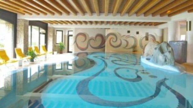 An indoor swimming pool with a blue and white design, wooden ceiling beams, and yellow lounge chairs. - Seiwald (Kirchdorf in Tirol)