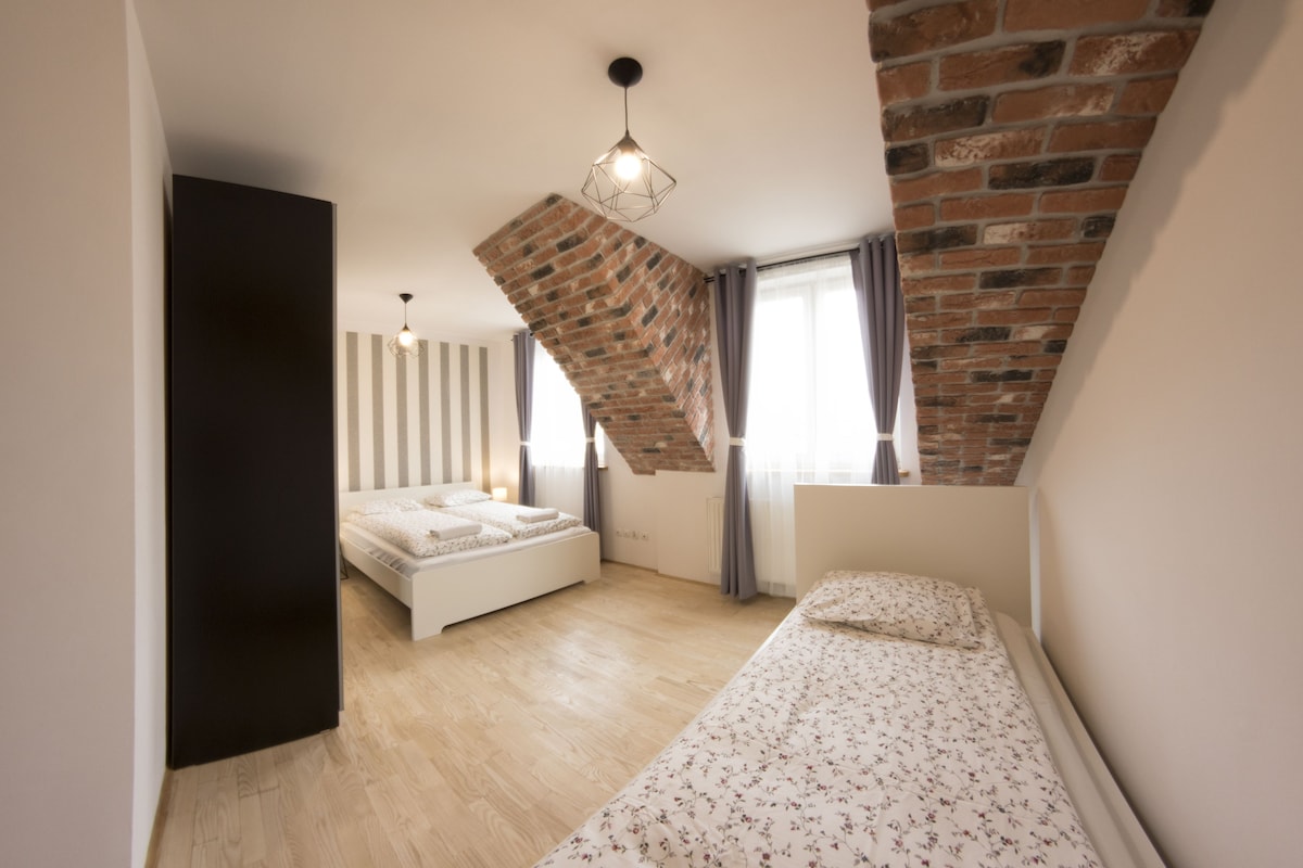 A modern bedroom with a loft bed, a small sofa, and a sloped ceiling with exposed brick.