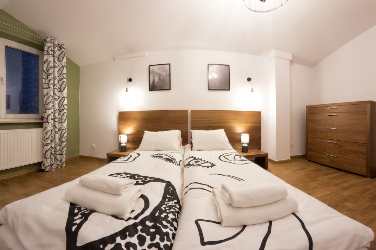 A neatly arranged bedroom with a double bed, two bedside tables with lamps, a wooden dresser, and a window with a curtain.