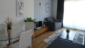 A modern living room with a glass dining table, a grey sofa, a flat-screen TV, and a clock on the wall.