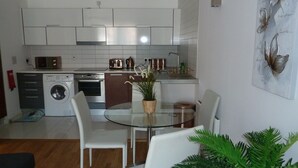 A compact kitchen with a washing machine, oven, and toaster. There is a glass dining table with chairs and a potted plant.