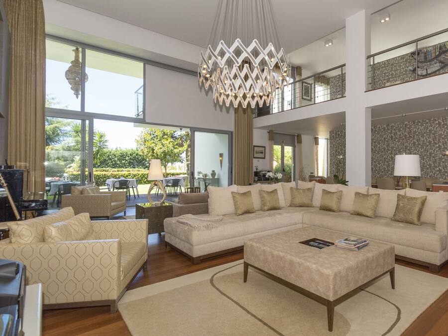 A spacious living area with a large sectional sofa, a coffee table, and a chandelier. There is a balcony with a railing and a view of the outdoors.