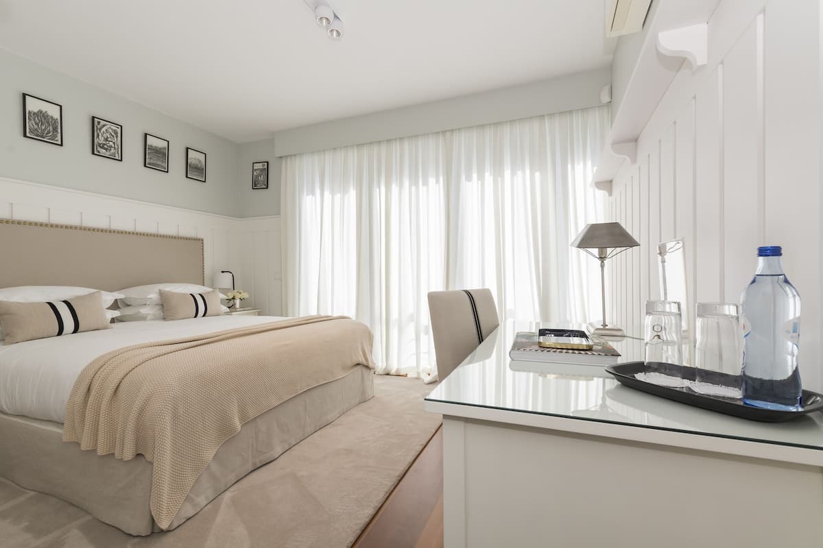 A modern hotel room with a large bed, a desk with a lamp, a chair, and a tray with a bottle and glasses.
