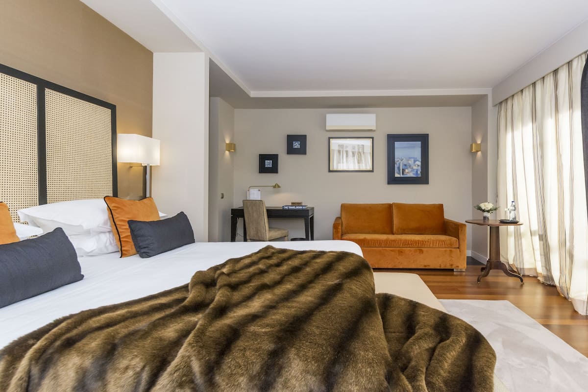 A hotel room with a large bed, an orange sofa, a desk with a lamp, and a view of the city through the window.