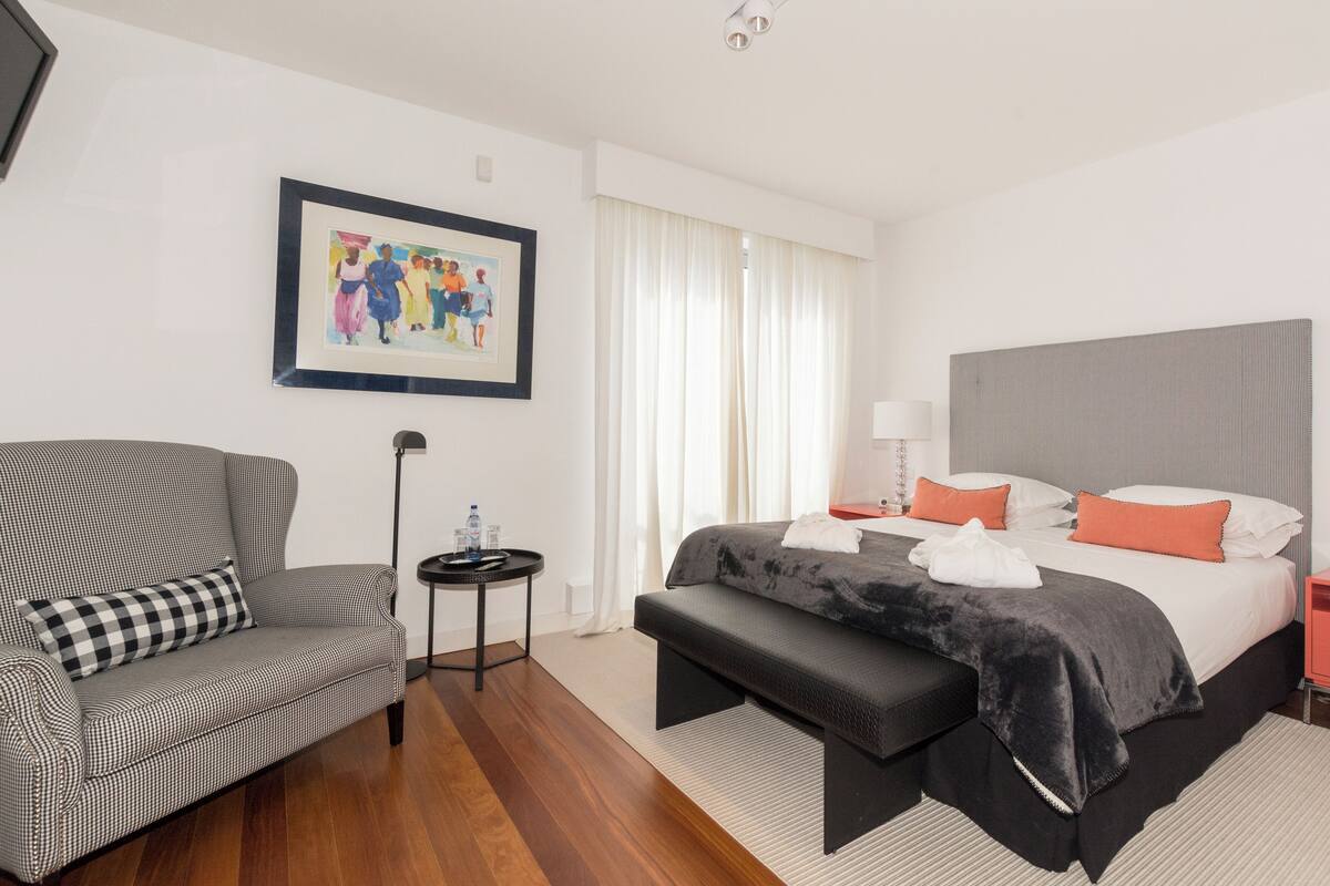A hotel room with a checkered sofa, a bed with a grey headboard, and a framed painting on the wall.