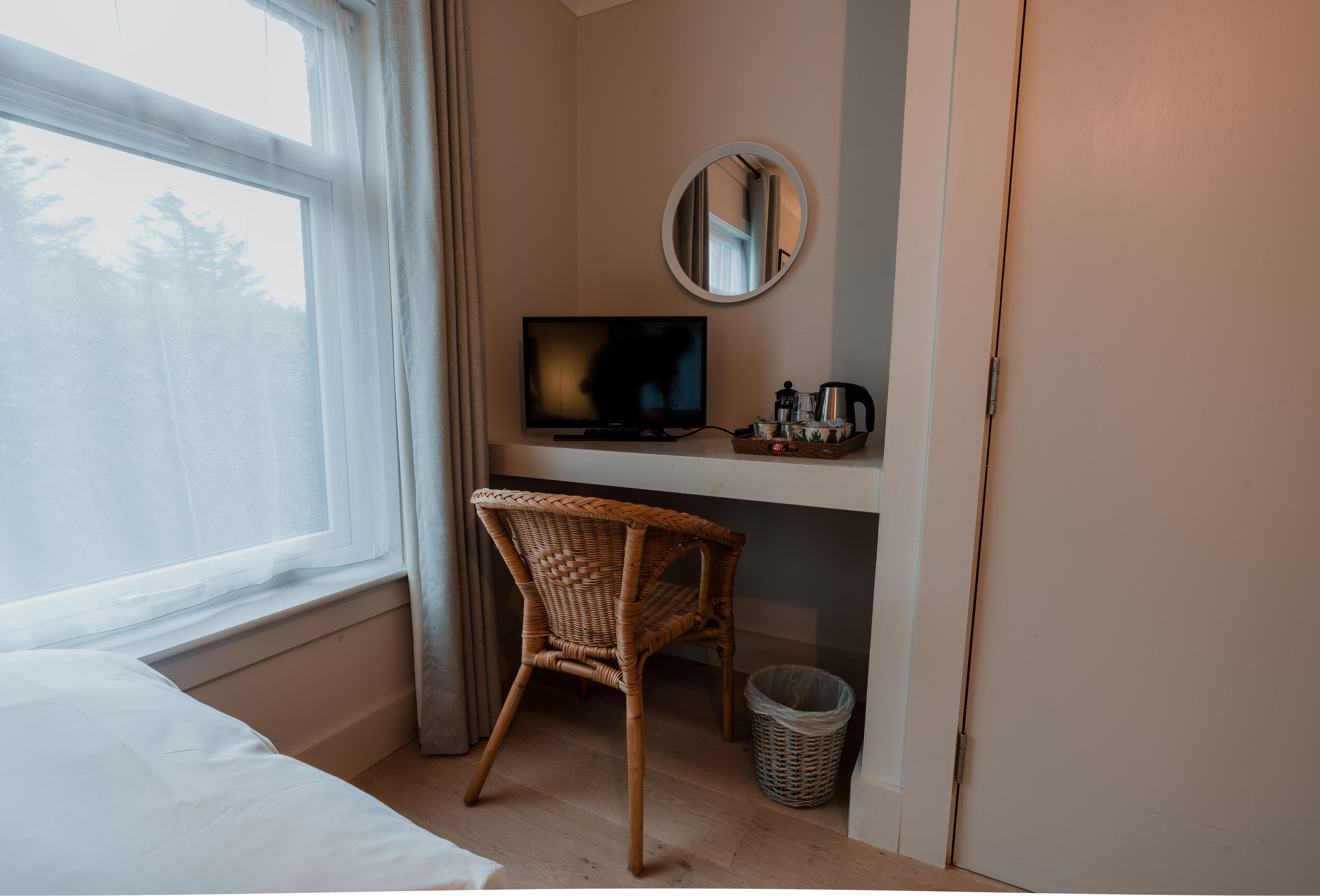 Quarto com uma cama, uma cadeira de vime, televisão, uma mesinha com chaleira e um cesto de lixo.
