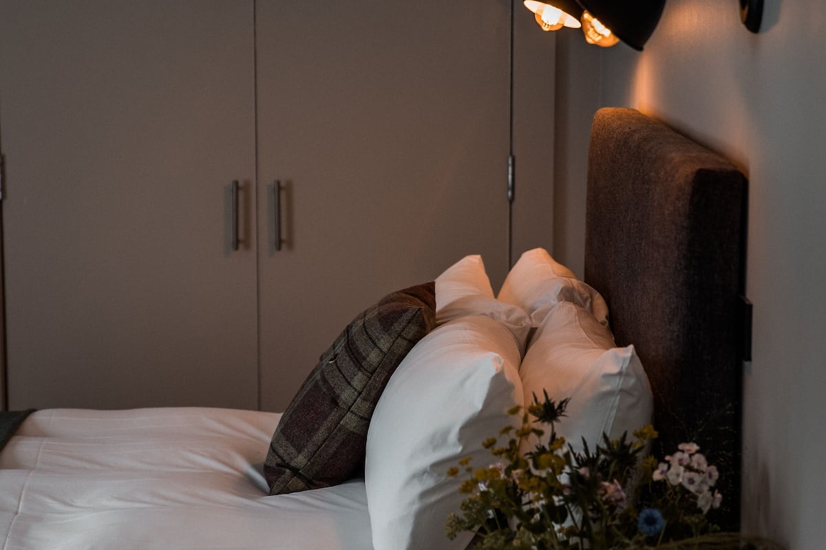 A bedroom with a bed, pillows, a bedside table with a vase of flowers, and a wall-mounted lamp.