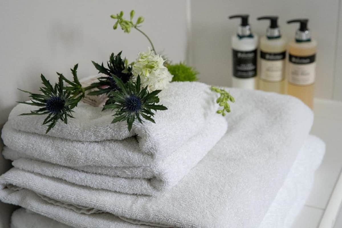 A stack of white towels with a sprig of greenery and a white flower on top, placed on a white surface. Behind them are three bottles of liquid soap.