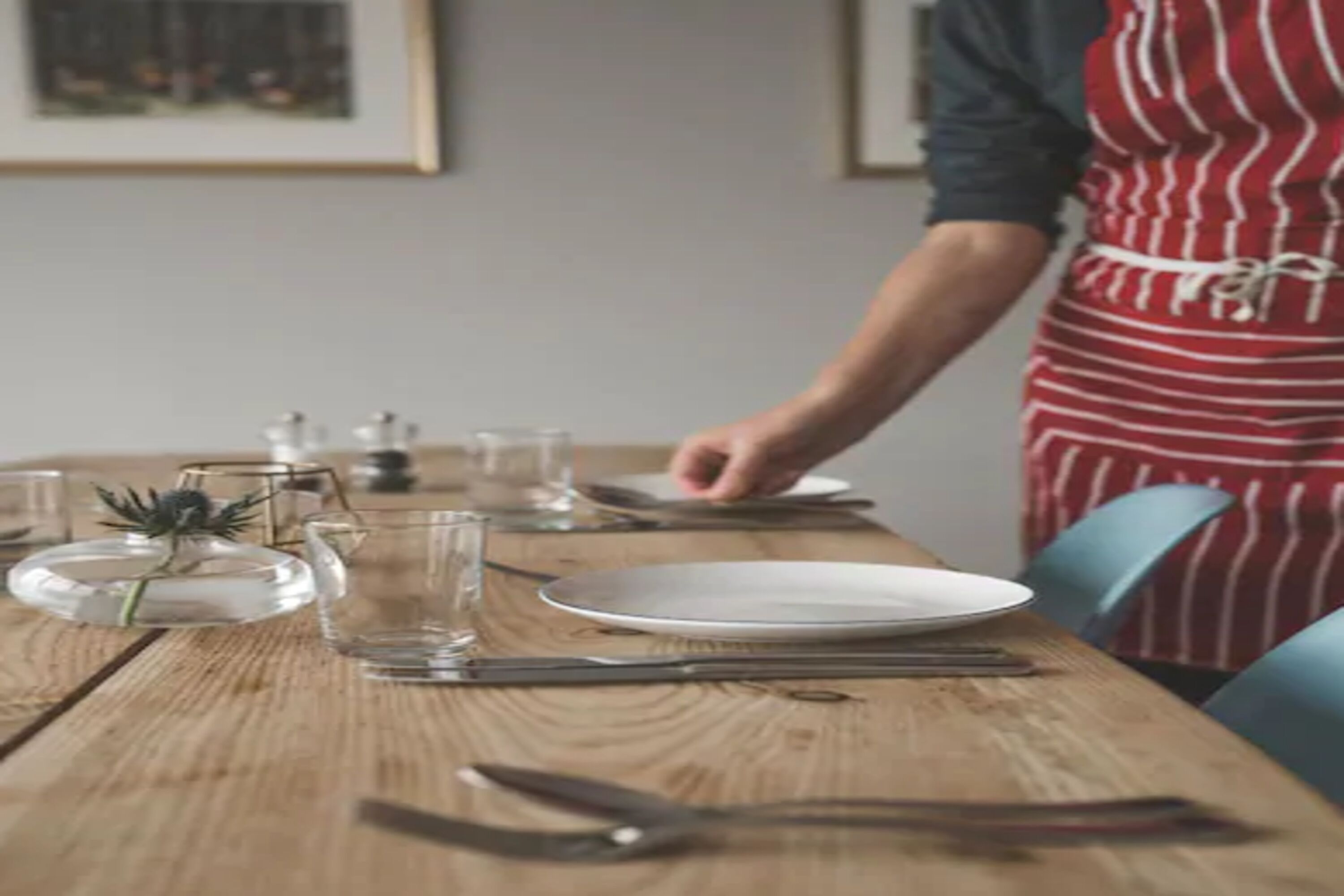 Una mesa de comedor puesta con platos, vasos y cubiertos, y una persona con delantal rojo y blanco a punto de servir.