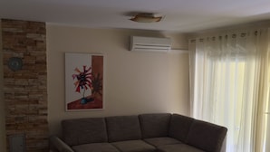 A living room with a sectional sofa, a coffee table, a stone accent wall, a painting, an air conditioning unit, and a window with curtains.