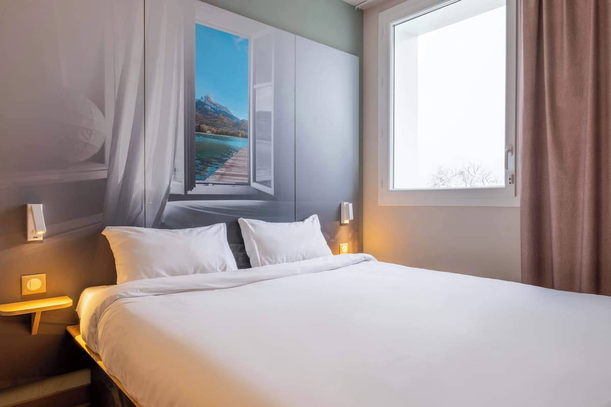 A hotel room with a large bed, a view of a lake and mountains through an open window, and a nightstand with a lamp.