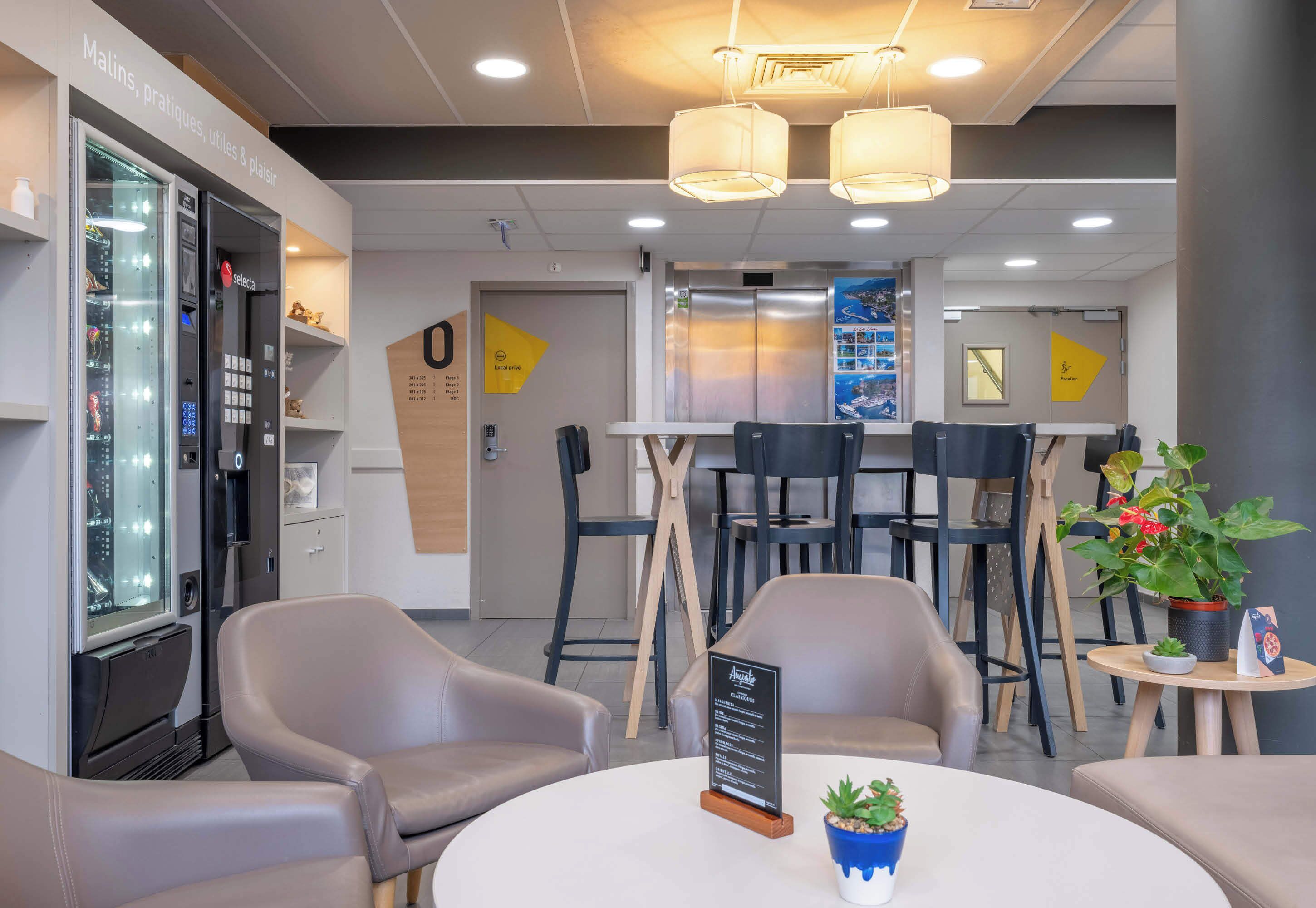 Een moderne hotel lobby met een zelfbedieningskiosk, een zithoek en een receptiebalie.