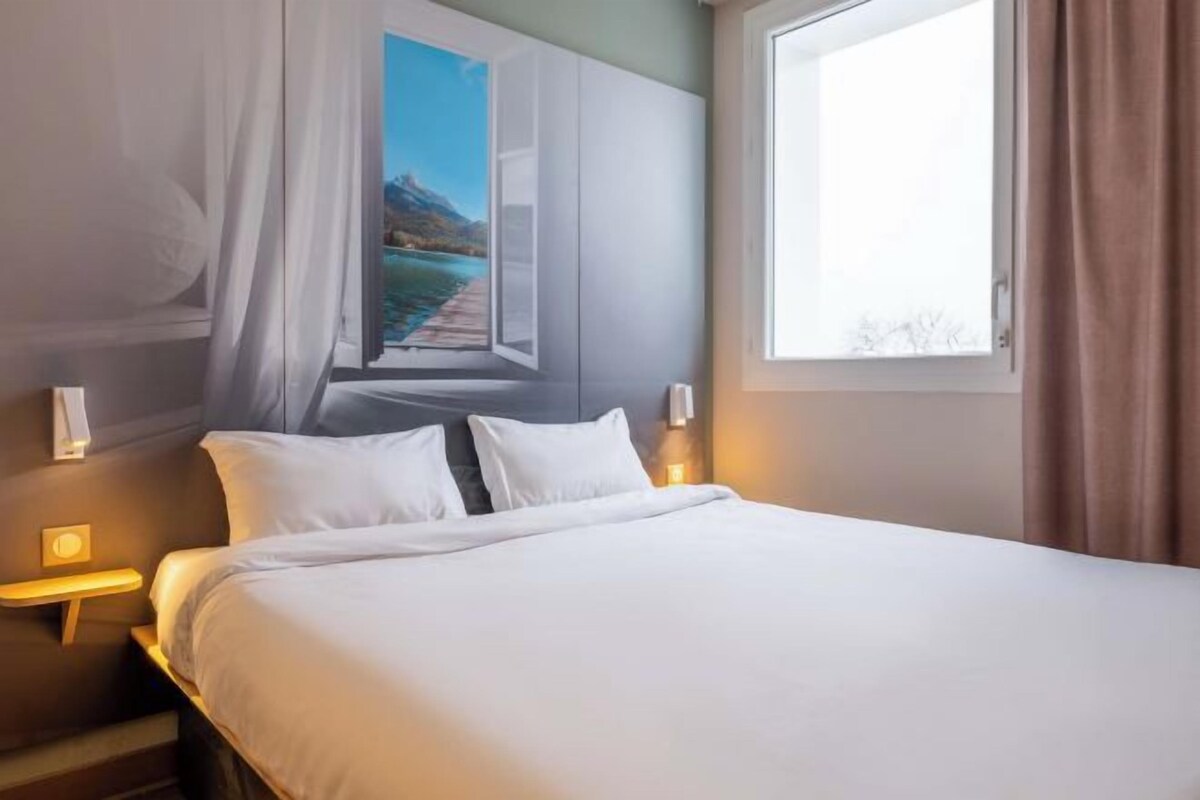 A hotel room with a large bed, a nightstand, a wall-mounted lamp, and a window with curtains.