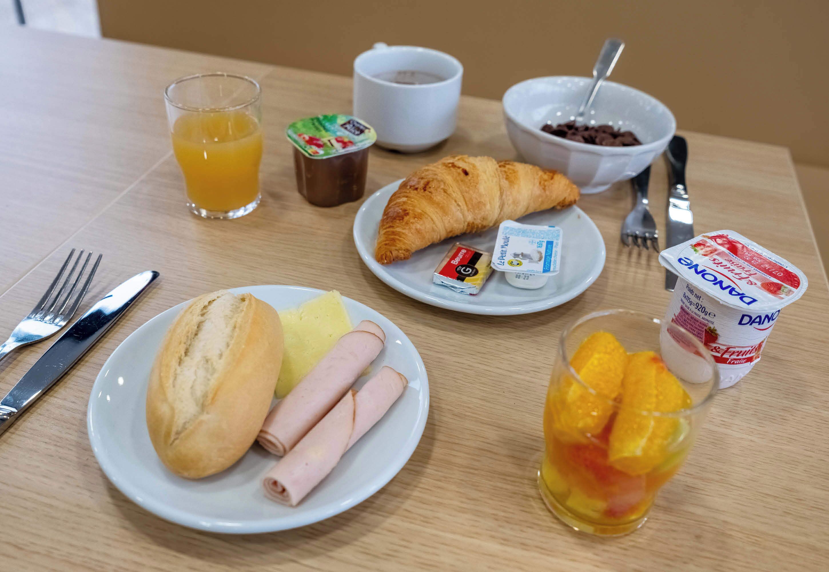 Una mesa de desayuno con cruasanes, jamón, pan, zumo de naranja, yogur y una taza de café.