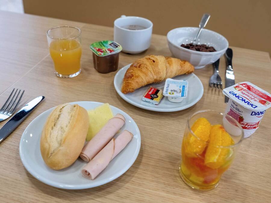 Una mesa de desayuno con cruasanes, jamón, pan, zumo de naranja, yogur y una taza de café.