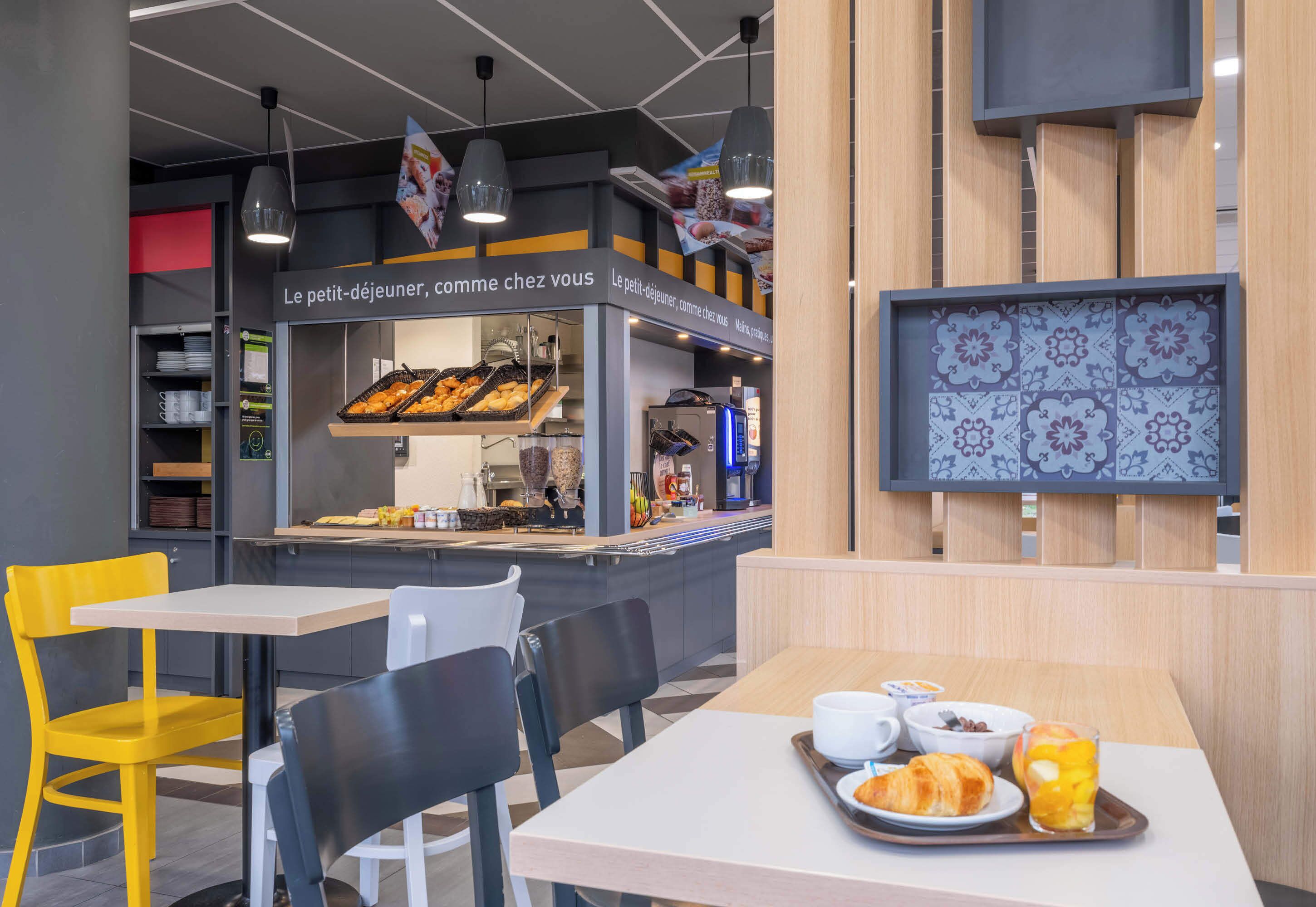 Ein Café mit einer Theke, auf der Gebäck und eine Kaffeemaschine präsentiert werden, Tische mit Stühlen und einer Tablett mit Croissants und Früchten.