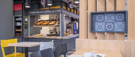 Ein Café mit einer Theke, auf der Gebäck und eine Kaffeemaschine präsentiert werden, Tische mit Stühlen und einer Tablett mit Croissants und Früchten.