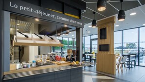 A modern cafe with a self-service counter, a large window offering a view of the outside, and a sign above the counter reading "Le petit-déjeuner, comme chez vous". - B&B HOTEL ANNEMASSE Saint-Cergues (Saint-Cergues)