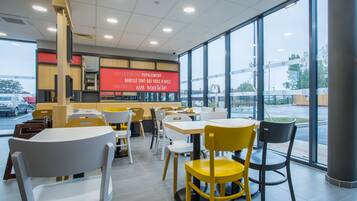 A modern restaurant interior with tables and chairs, a large window with a view of the outside, and a red sign with white text.