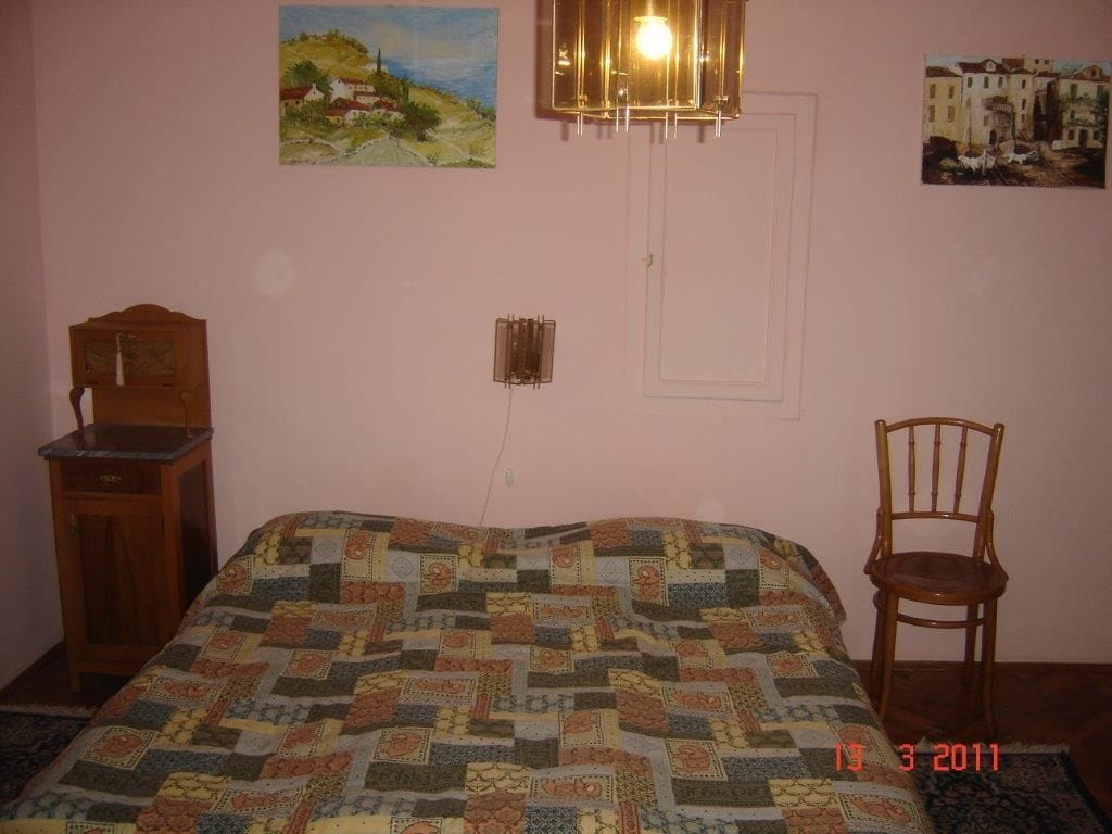 A bedroom with a quilted bedspread, a wooden cabinet, a chair, and two paintings on the wall.