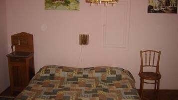 A bedroom with a quilted bedspread, a wooden cabinet, a chair, and two paintings on the wall.