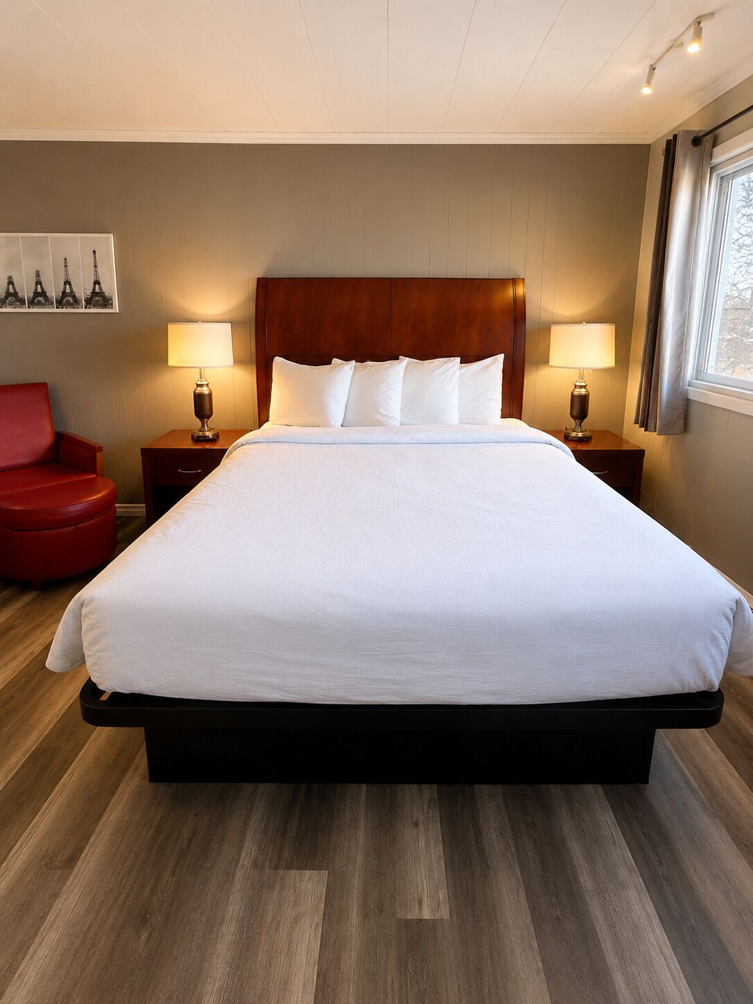 A large bed with a wooden headboard, two bedside tables with lamps, a red chair, and a window with curtains.