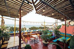 An outdoor seating area with bamboo roofs, tables, and chairs, overlooking the beach.