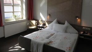 A hotel room with a large bed, a chair, a small table, and a window with curtains.