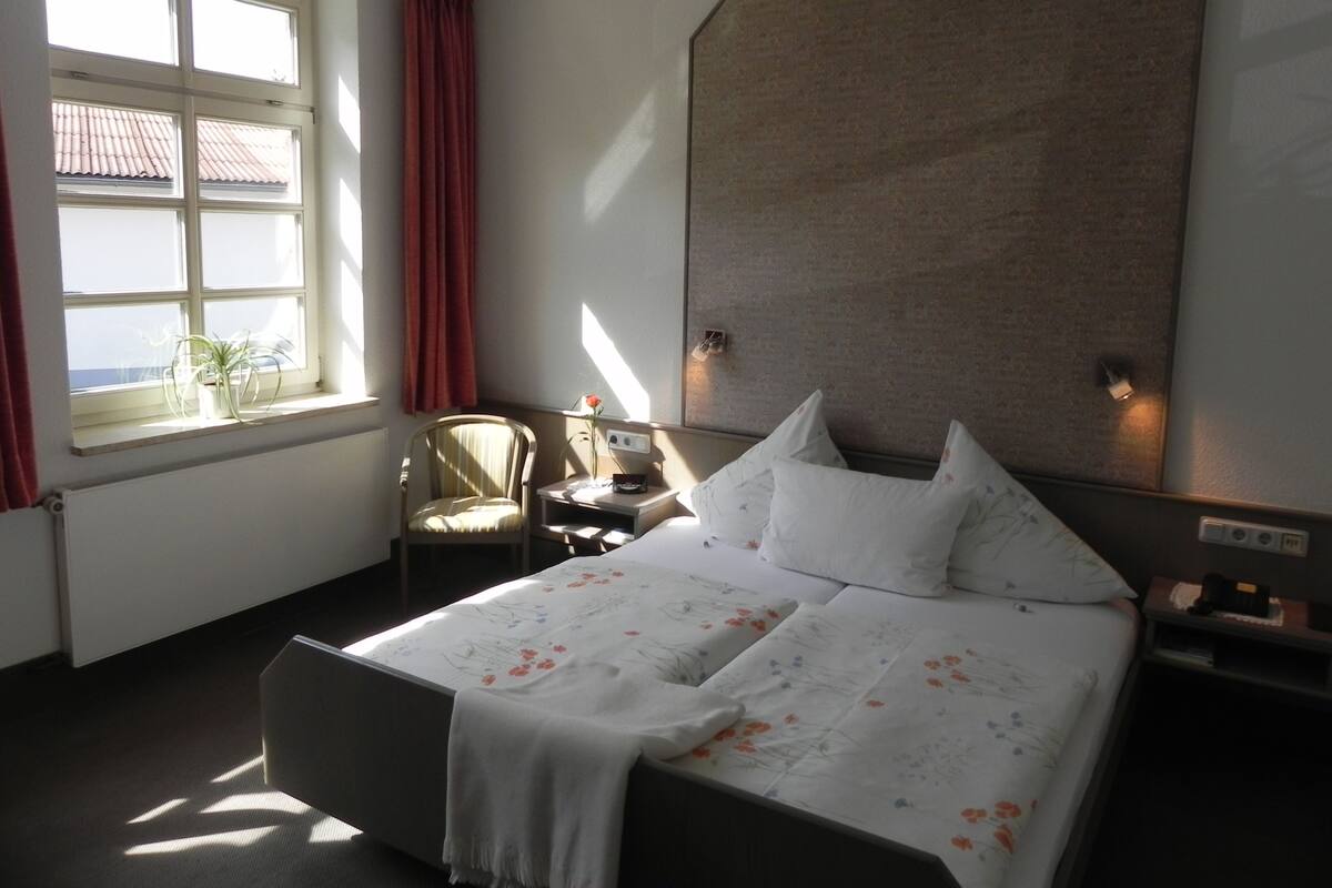 A hotel room with a large bed, a chair, a small table, and a window with curtains.