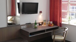 A hotel room with a TV mounted on the wall, a desk with a basket of fruit, a chair, and a window with red curtains.