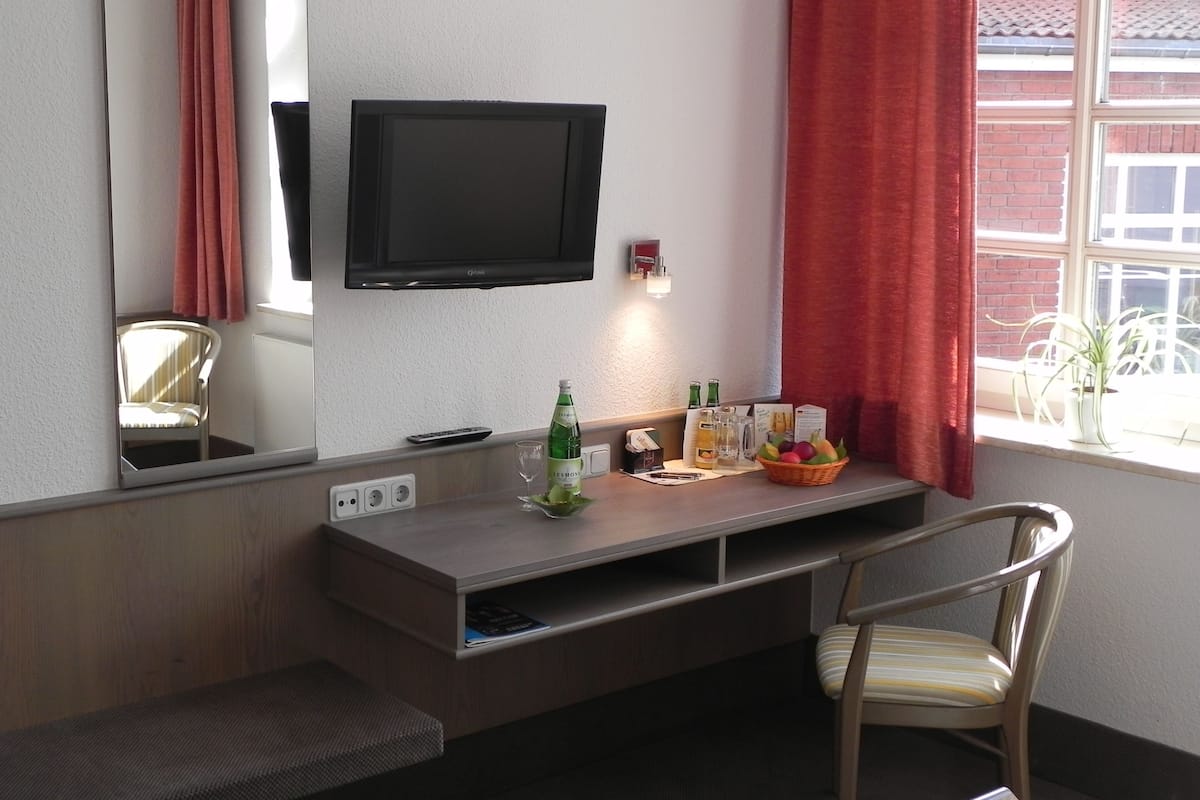 A hotel room with a TV mounted on the wall, a desk with a basket of fruit, a chair, and a window with red curtains.