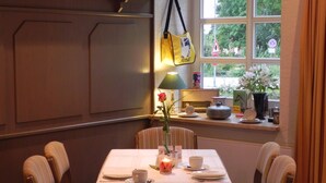 A dining area with a table set for two, chairs, a window with a view of greenery, and a lamp on the table.