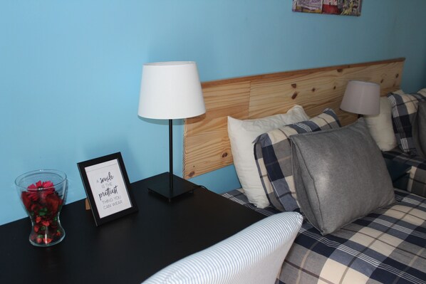 A neatly made bed with plaid bedding, a bedside table with a lamp, a framed inspirational quote, and a vase with red flowers. - TripGeo Guesthouse (Lisbon)