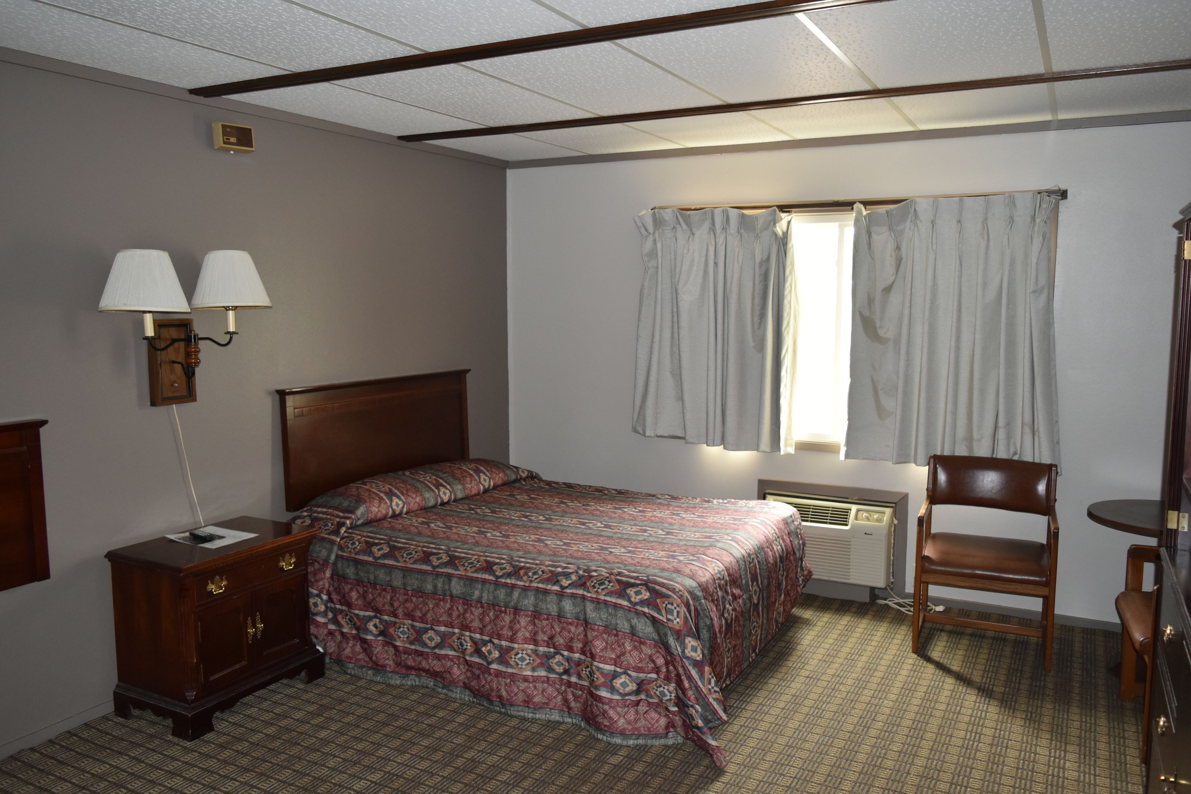 Una habitación de hotel con una cama, una mesita de noche, una silla, una lámpara y una ventana con cortinas.