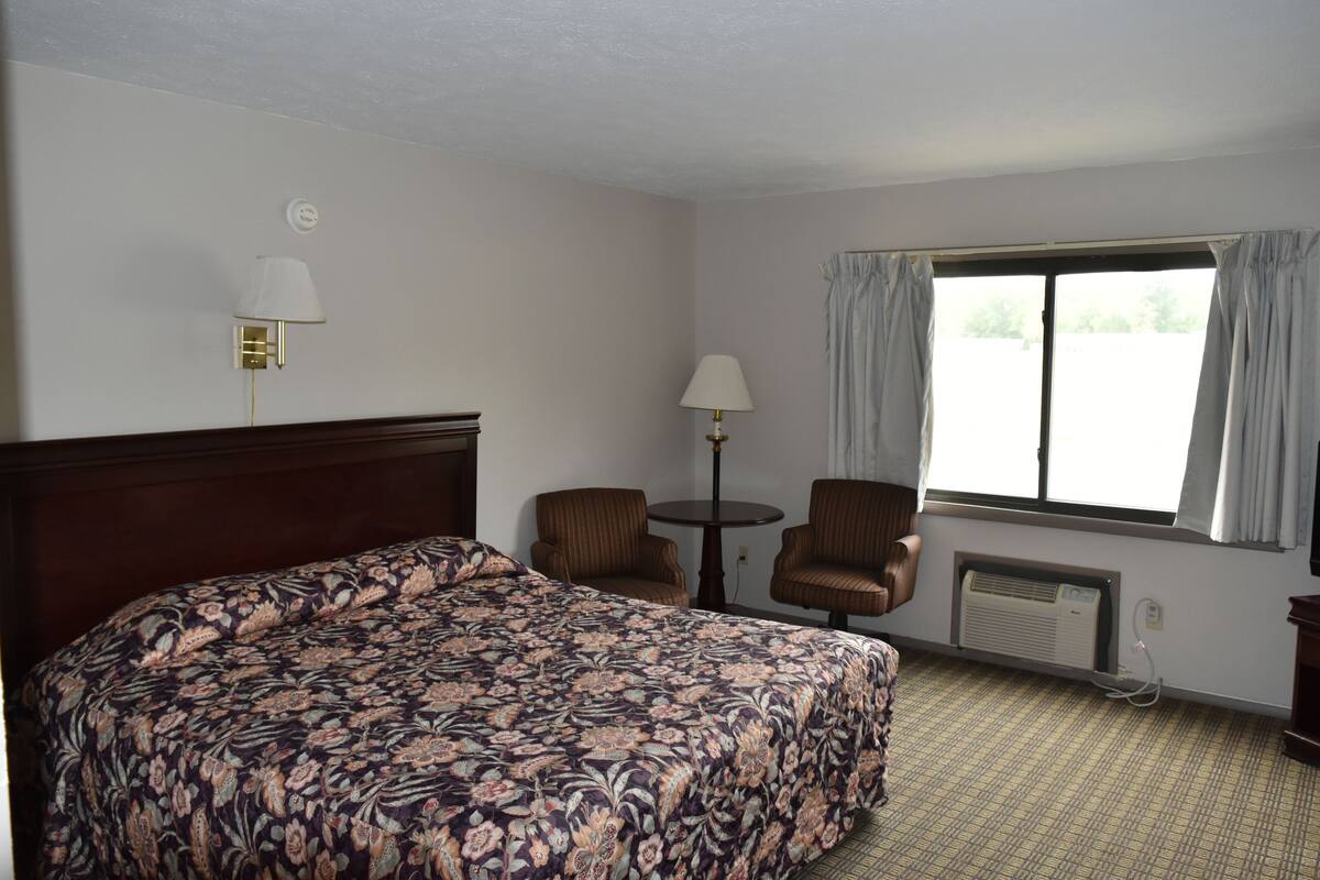 A hotel room with a large bed, two armchairs, a small table, a window with curtains, and a wall-mounted air conditioner.