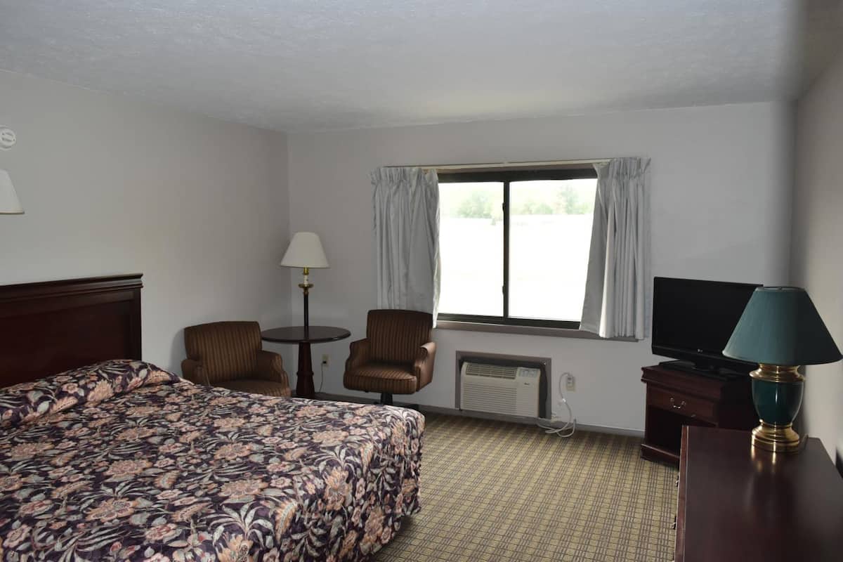 A hotel room with a bed, a chair, a small table, a lamp, a television, and a window with curtains.