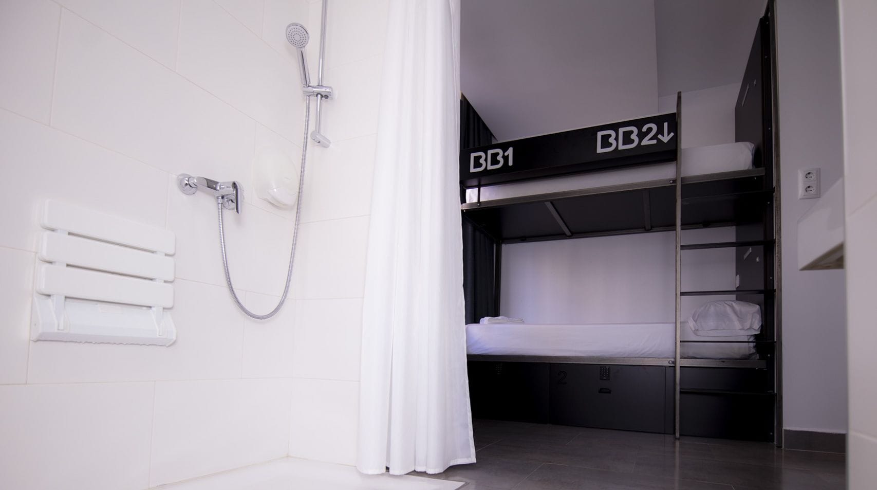 Une chambre d’hôtel compacte équipée d’une douche, d’un porte-serviettes et de deux lits superposés étiquetés BB1 et BB2.