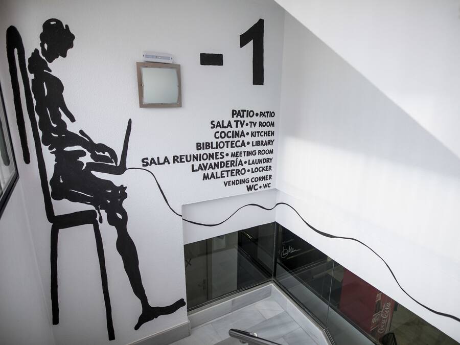 Un interior moderno con la silueta negra de una persona sentada en una silla, un letrero en la pared que enumera nombres de habitaciones y funciones, y una barandilla de cristal.