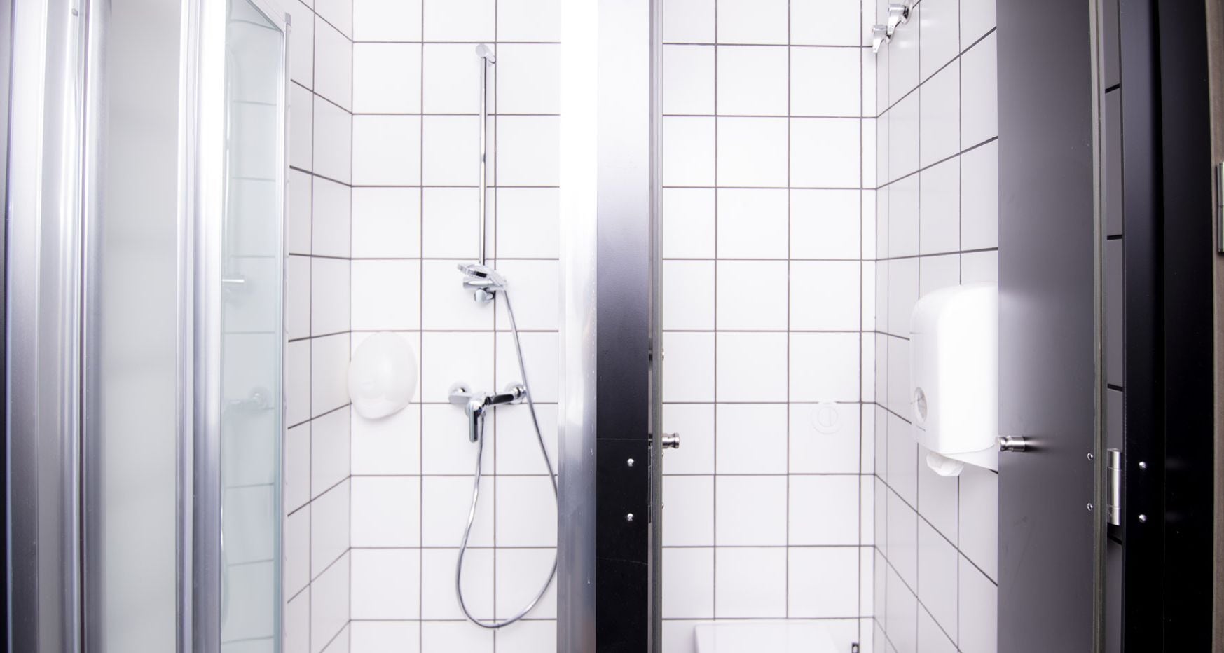Un baño moderno con paredes de azulejos blancos, cabina de ducha con mampara de vidrio, cabezal de ducha de mano y dispensador de toallas de papel.