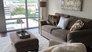 A modern living room with a large window, a sofa with patterned cushions, a fluffy white throw, and a side table with a lamp. - New World Stay (McLean)